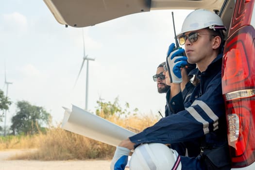 Close up technician worker man use walkie talkie to contact his co-worker and sit in the back of van in area of power plant with wind tubine or windmill.