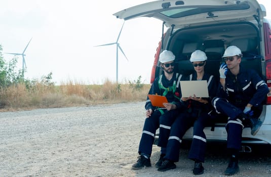 Group of technician workers sit on back of van on the road and discuss about work using laptop with windmill or wind turbine on the background.