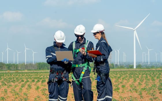 Close up professional technician workers man and woman stand in front of row of windmill or wind turbine and work together using laptop with day light and they look happy with smiling.