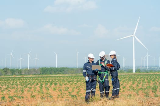 Side view technician workers man and woman stand in front of row of windmill or wind turbine and work together using laptop with day light.