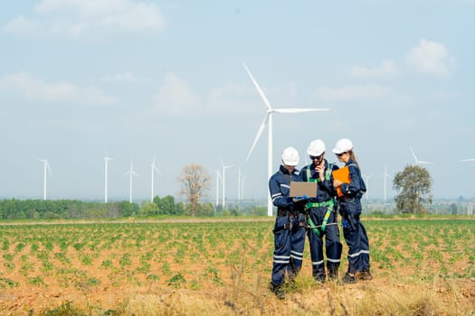 Wide shot technician workers man and woman stand in front of row of windmill or wind turbine and work together using laptop with day light.