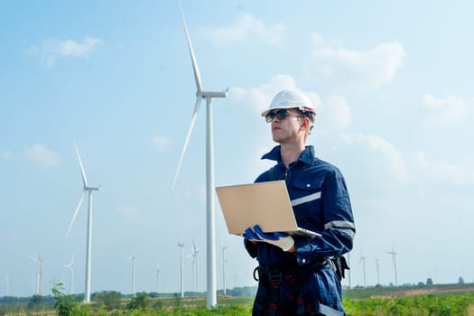 Portrait of technician worker hold laptop and look to left side in front of windmill or wind turbine with blue skay in area of power plant factory.