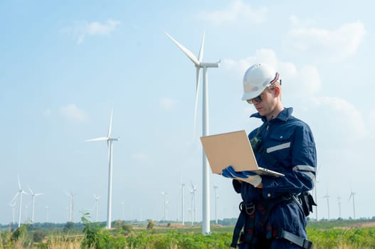 Technician worker hold laptop and look to the monitor for working in front of windmill or wind turbine with blue skay in area of power plant factory.