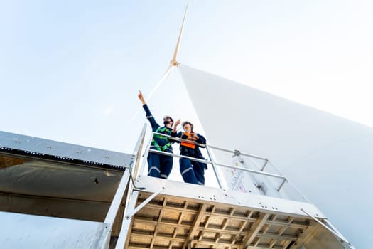 Lower view of professional technician man and woman stay on base of windmill or wind turbine and man also point to the left with blue sky.