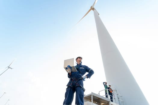 Wide shot professional technician man stand with hold laptop and stand near base of windmill or wind turbine also look to right side with his co-worker in the back.