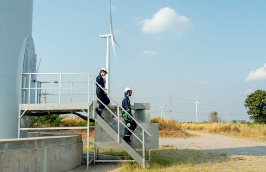 Two of technician workers walk down from stair of base of wind turbine or windmill after finish maintenance work in area of power plant.