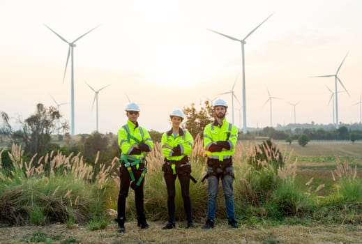 Group of technician workers stand with arm crossed and look at camera in front of windmill or wind turbine and evening light in area of power plant.