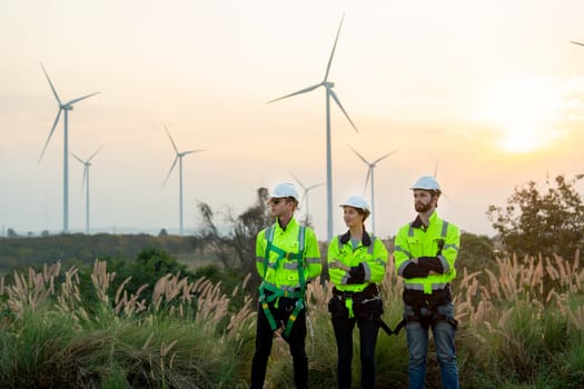 Group of technician workers stand with arm crossed and look to left side in front of windmill or wind turbine and evening light in area of power plant.