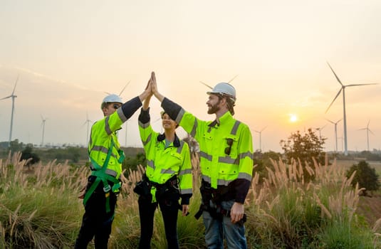 Group of technician worker man and woman stand with touch hands together express happiness during work and stand in front of windmill or wind turbine farm in area of power plant.
