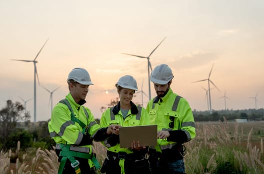 Group of professional technician workers man and woman work using laptop and stay in front of windmill or wind turbine with evening light.