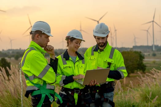 Close up group of technician workers man and woman work using laptop and stay in front of windmill or wind turbine with evening light.