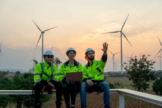 Professional technician workers with safety suit sit and discuss work using laptop and row of windmill or wind turbine on the background and evening light.