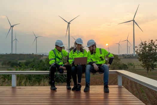 Group of professional technician workers with safety suit sit and discuss work using laptop and row of windmill or wind turbine on the background and evening light.