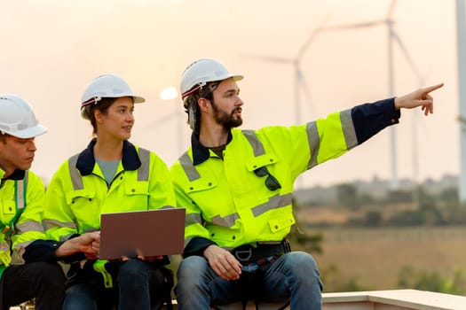 Close up technician worker man point to right side and stay with his co-worker using laptop to discuss work in front of windmill or wind turbine with evening light.