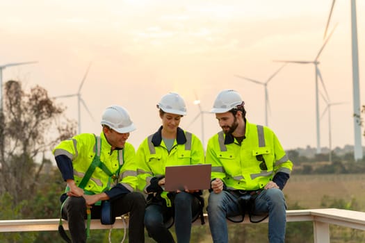 Group of professional technician workers with safety suit sit and discuss work using laptop and row of windmill or wind turbine on the background in evening.