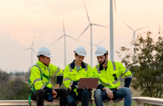 Professional technician workers with safety suit sit and discuss work using laptop and row of windmill or wind turbine on the background in evening.