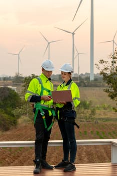 Vertical image professional technician workers stand together in front of windmill or wind turbine using laptop to discuss about work with evening light.