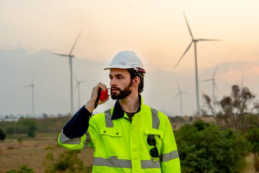 Portrait technician worker man use walkie talkie to contact his co-worker in evening with windmill or wind turbine on the background.