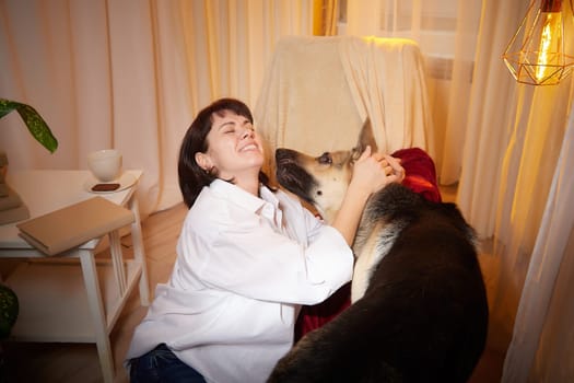 Adult mature woman with big shepherd dog in white shirt. Room with girl and calm cozy evening atmosphere with transparent curtains and soft warm light of lamps. Concept of love for animals, pets