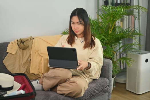 Pleasant young woman booking flight tickets on digital tablet and packing suitcase in living room.