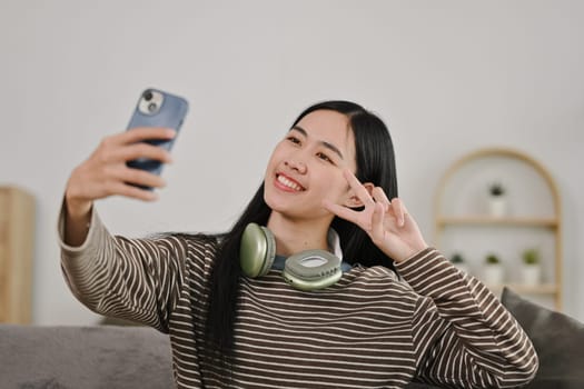 Cheerful Asian woman showing two fingers near her face taking selfie on smartphone at home.