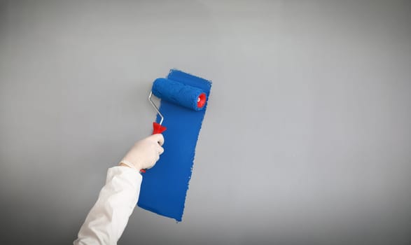 Male painter in uniform paints wall with roller. Professional uses simple hand tool. Paint large areas in a short time. Uniform distribution color and end result. Scratches during painting