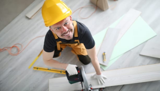 Smiling carpenter in helmet sawing laminate flooring. Simple and popular layout option. Protective varnish quality, density and strength base strip. Brand awareness and value material