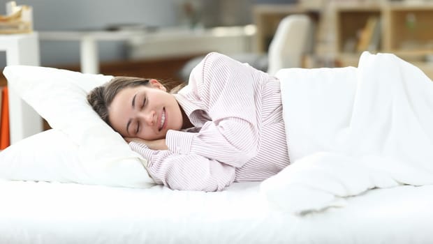 Happy girl in pajamas lies in bed closing her eyes. Woman is disturbed by insomnia, normalization sleep. Girl sleeps at home under white blanket. Healthy sleep during self-isolation
