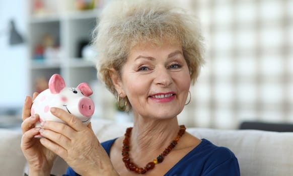 Happy elderly woman in apartment shows piggy bank. Accumulation pension capital. Elderly woman happily saving money in piggy bank. Older generation is used to saving and saving money