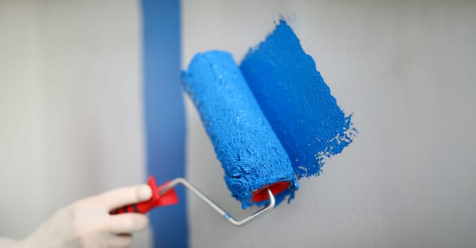 Hand holds roller and wire on wall with blue paint. Low roller performance. Convenient and versatile tool for painting walls. Tools with which best lay paint on surface. DIY repair in apartment