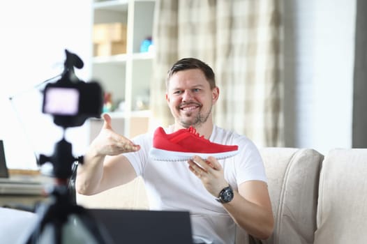 Man shows red sneaker in front camera, video blog. Quality product description. Guy works remotely, offering goods online. Man in his apartment takes an overview new sports shoes