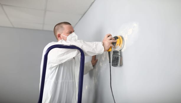 Male builder in uniform cleans wall with putty. Quality putty surfaces. Man removes an old putty with an electric tool. Repair walls, alignment before painting. Finishing putty walls apartment