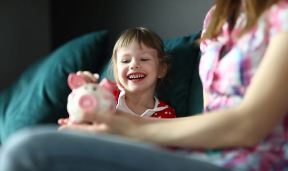 Little girl laughs and puts coin in piggy bank. Impact pandemic on economy and financial situation families. Personal cost accounting. Teach child how to save money. Financial literacy