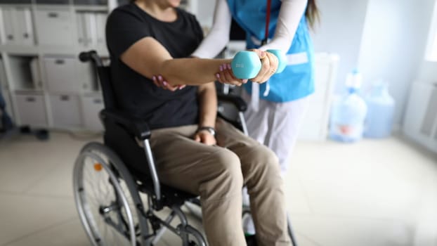 Man on wheelchair works out his hand with dumbbell. Simple exercises for disabled. Doctor helps restore health. Exercise is suitable for people with disabilities who have power in their upper body