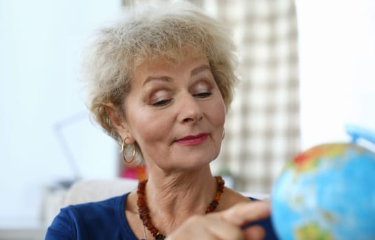 Elderly woman in apartment shows finger on globe. Search for old friends or relatives with whom you lost contact. Desire to travel in old age. Woman dreams rest around world during quarantine