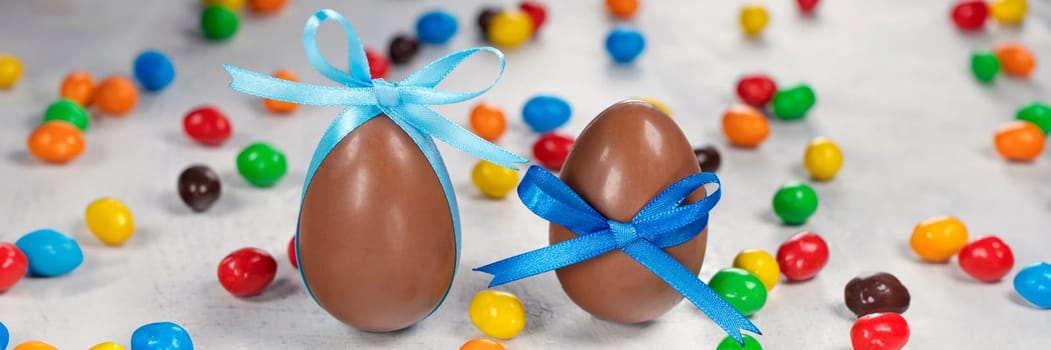 banner of two big chocolate eggs with a blue bow on a white concrete background with colorful little eggs. Concept of Happy Easter. Soft focus