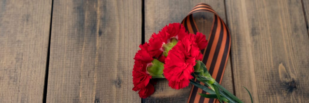 banner of three red carnations with a St. George ribbon on a wooden background. Georgievsk ribbon - Russian symbol of victory and three red carnations. Holiday card for the holiday of May 9. Soft focus