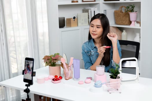 Young beautiful Asian showing cosmetic makeup lipstick matte pastel color testing in bestseller collection on arm test promotion on social media online recording smartphone at modern room. Stratagem.