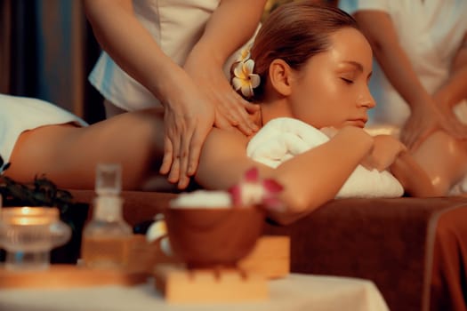 Caucasian couple customer enjoying relaxing anti-stress spa massage and pampering with beauty skin recreation leisure in warm candle lighting ambient salon spa at luxury resort or hotel. Quiescent