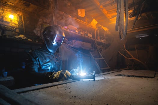 A welder in a workshop welds a metal part. General plan of an old cluttered garage where a man in protective gloves and a mask is making.