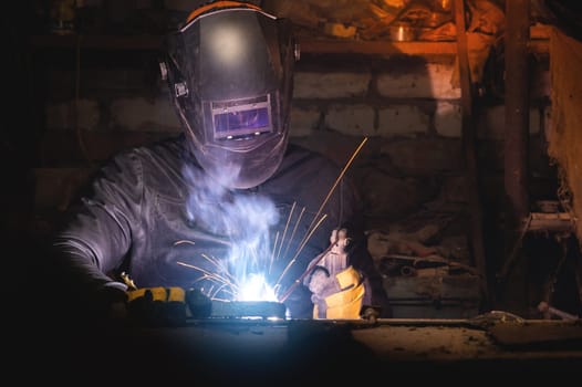 Village workshop, welding in an old garage. An unrecognizable man repairs a metal part wearing protective gloves and a mask.