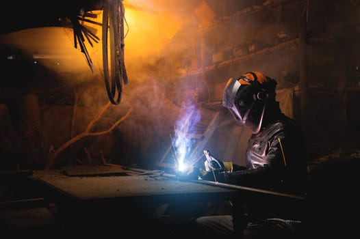 Village workshop, welding in an old garage. An unrecognizable man repairs a metal part wearing protective gloves and a mask.