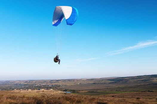 A paraglider takes off from a mountainside with a blue and white canopy and the sun behind. A paraglider is a silhouette. The glider is sharp, with little wing movement. A male paraglider launches a paraglider into the air.