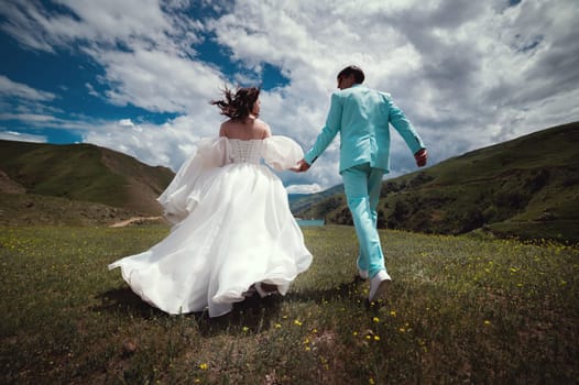 Rear view of running loving newlyweds holding hands, a bride in a wedding dress and a wife in a turquoise suit running across a field against the backdrop of green hills.