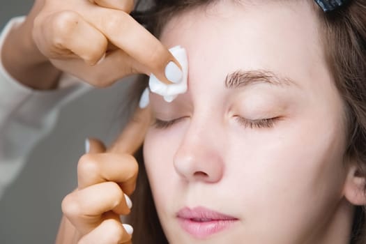 In a beauty salon, a master cosmetologist applies makeup and wipes eyebrows with a cotton pad. Close-up of the model's face during the procedure.