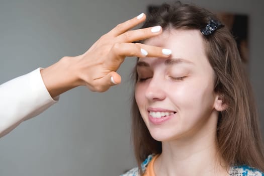 In a beauty salon, a master cosmetologist applies makeup and wipes eyebrows with a cotton pad. Close-up of the model's face during the procedure.