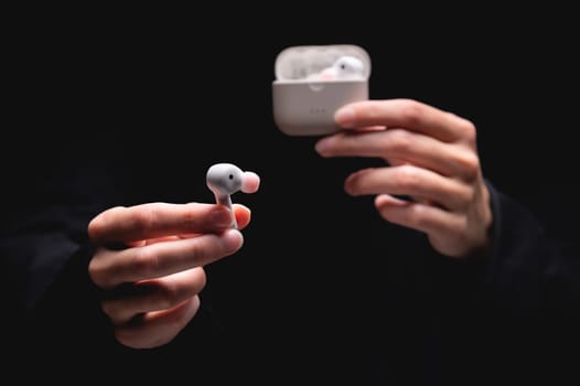 close-up of a woman taking a white wireless earphone out of the charger. Female hands touch the headphones of a portable gadget and present it forward to the camera on a black background.