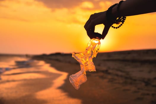 Volunteer and plastic bottle, collecting waste on the sea beach, pollution and recycling concept. hand holds a transparent plastic bottle in water lying on a sandy beach during sunset or dawn.