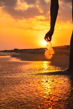 Volunteer and plastic bottle, collecting waste on the sea beach, pollution and recycling concept. hand holds a transparent plastic bottle in water lying on a sandy beach during sunset or dawn.