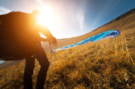 The paraglider is preparing to take off. A colorful kite lies on the grass, in the background there is a beautiful landscape, clear blue sky.